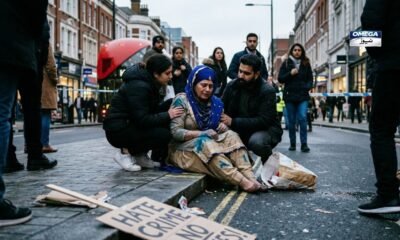 A photo of a sad Sikh woman sitting on a sidewalk in London. She wears a beige traditional outfit and a blue headscarf, both stained with blue powder. A young man and woman kneel beside her, offering comfort. People watch from the background behind blue police tape, and a cardboard sign on the ground reads "HATE CRIME."