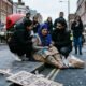 A photo of a sad Sikh woman sitting on a sidewalk in London. She wears a beige traditional outfit and a blue headscarf, both stained with blue powder. A young man and woman kneel beside her, offering comfort. People watch from the background behind blue police tape, and a cardboard sign on the ground reads "HATE CRIME."